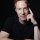 A man with light skin, short curly hair, and blue eyes is wearing a black t-shirt. He is resting his head on his finger, with his arm on a table, and has a calm expression, photographed against a dark background.