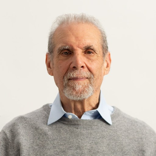 A mature person with a gray beard and mustache, wearing a light blue collared shirt under a gray sweater, is standing against a plain white background.