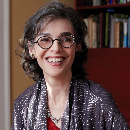 A woman with glasses and curly hair, smiling, wearing a sequined shawl and a red top, seated in a room with a bookshelf in the background.