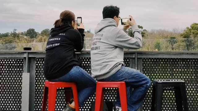 A man and a woman sitting on stools taking pictures with their phones.