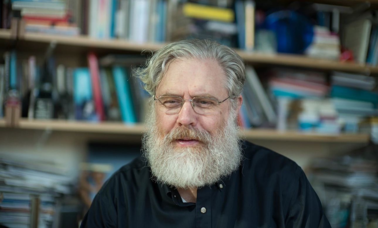 a man with a white beard and glasses sitting in front of a bookshelf.