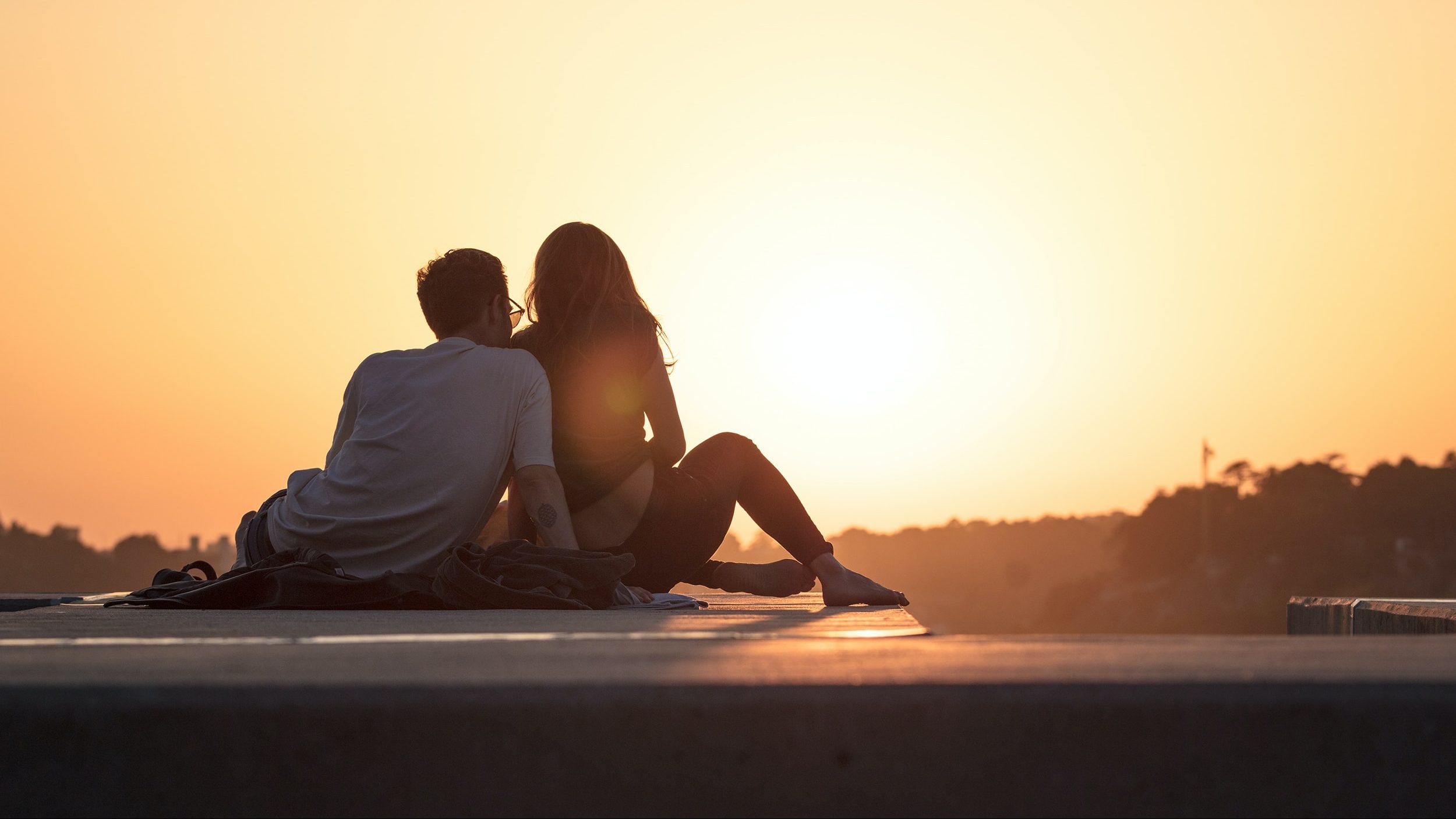 A man and a woman sitting on a ledge at sunset.
