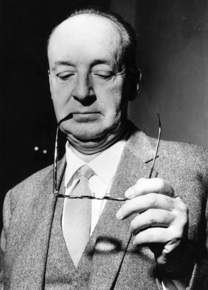 A darkly humorous man in a suit and tie brandishing a pair of glasses.