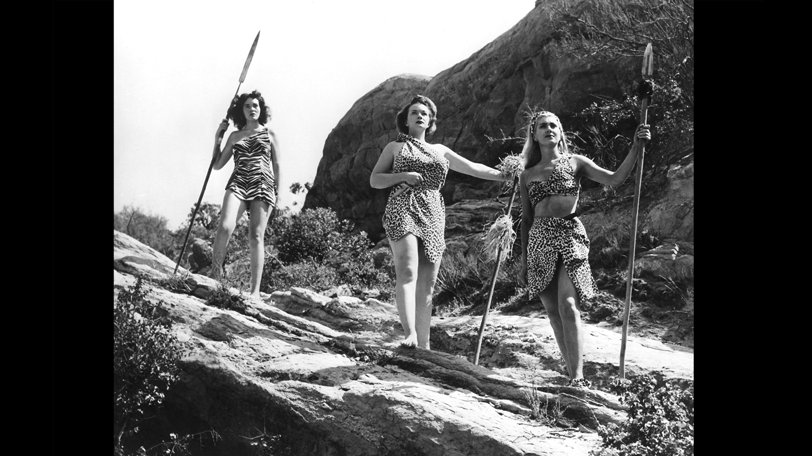 Three women, embodying the male hunter myth, standing confidently on a rock and armed with spears.