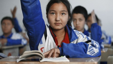 A girl in a blue jacket raises her hand in class, demonstrating language proficiency and recall.