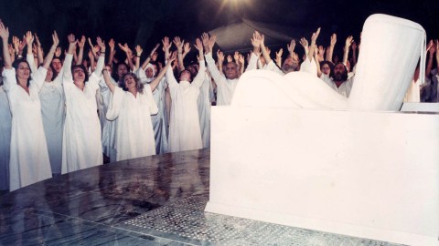 A group of people, part of a doomsday cult, in white robes standing in front of a white chair.