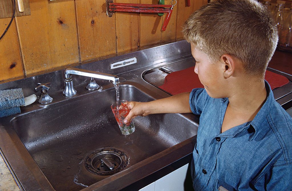 A boy is filling a glass with water in a kitchen sink.