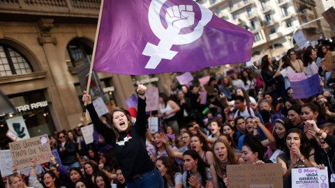 A group of feminists holding a purple flag in front of a crowd.