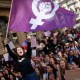 A group of feminists holding a purple flag in front of a crowd.