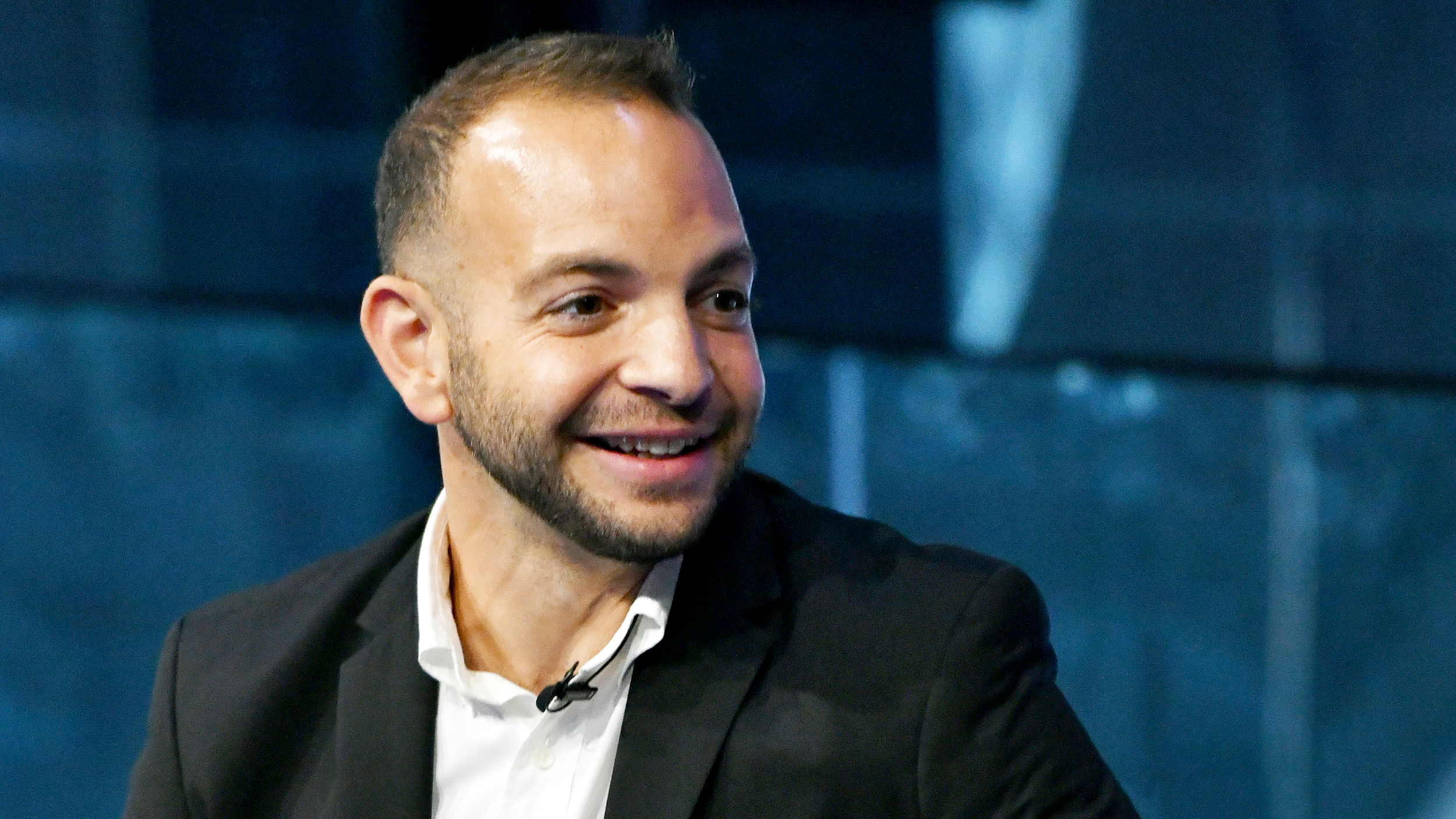 A man in a black suit and white shirt is smiling, unaffected by the doomer mindset.