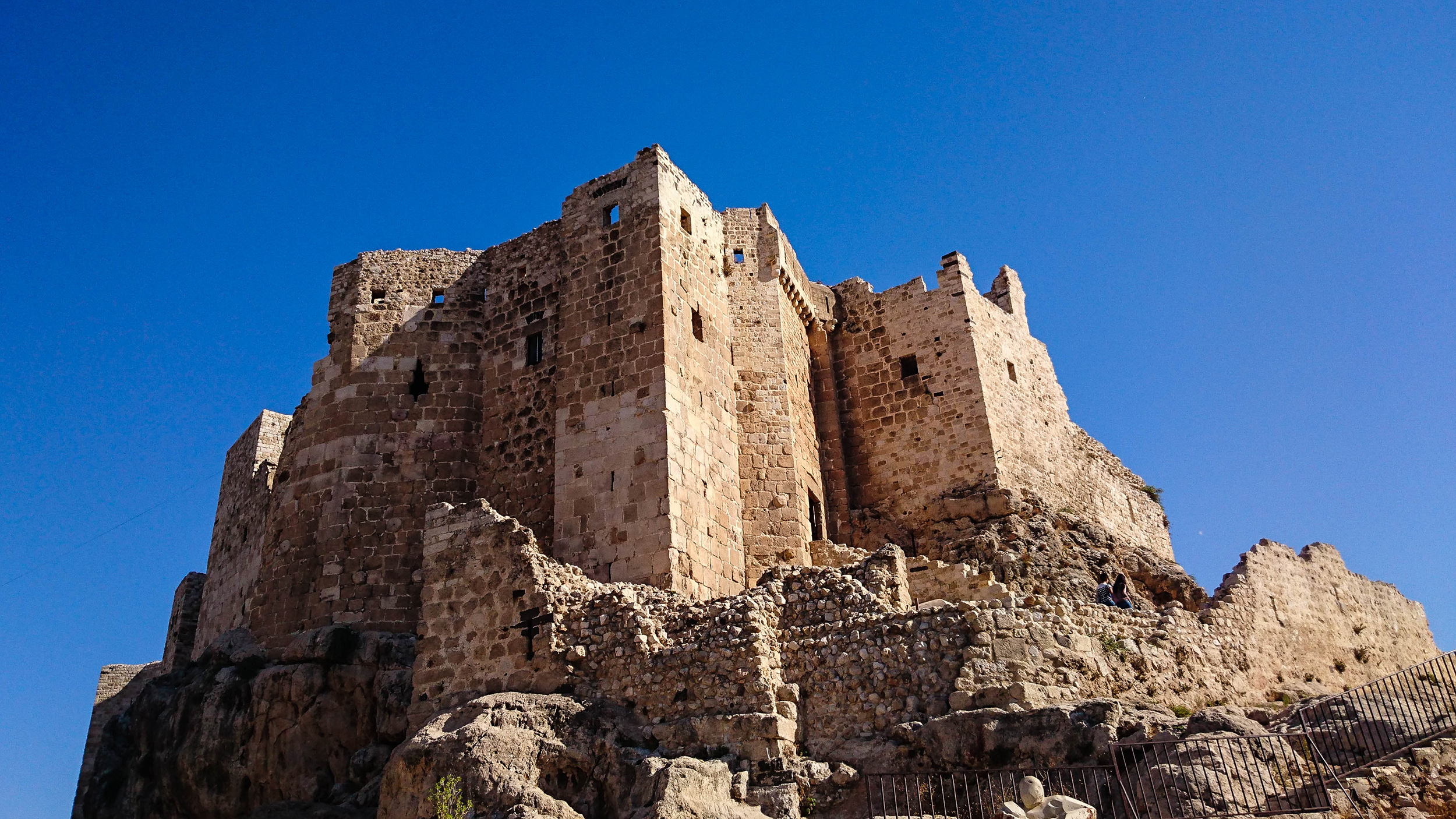 An ancient castle perches majestically atop a towering rock, under the expanse of a vivid blue sky.