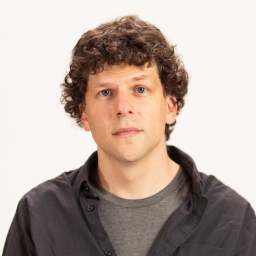 Jesse Eisenberg with curly hair is standing in front of a white background.