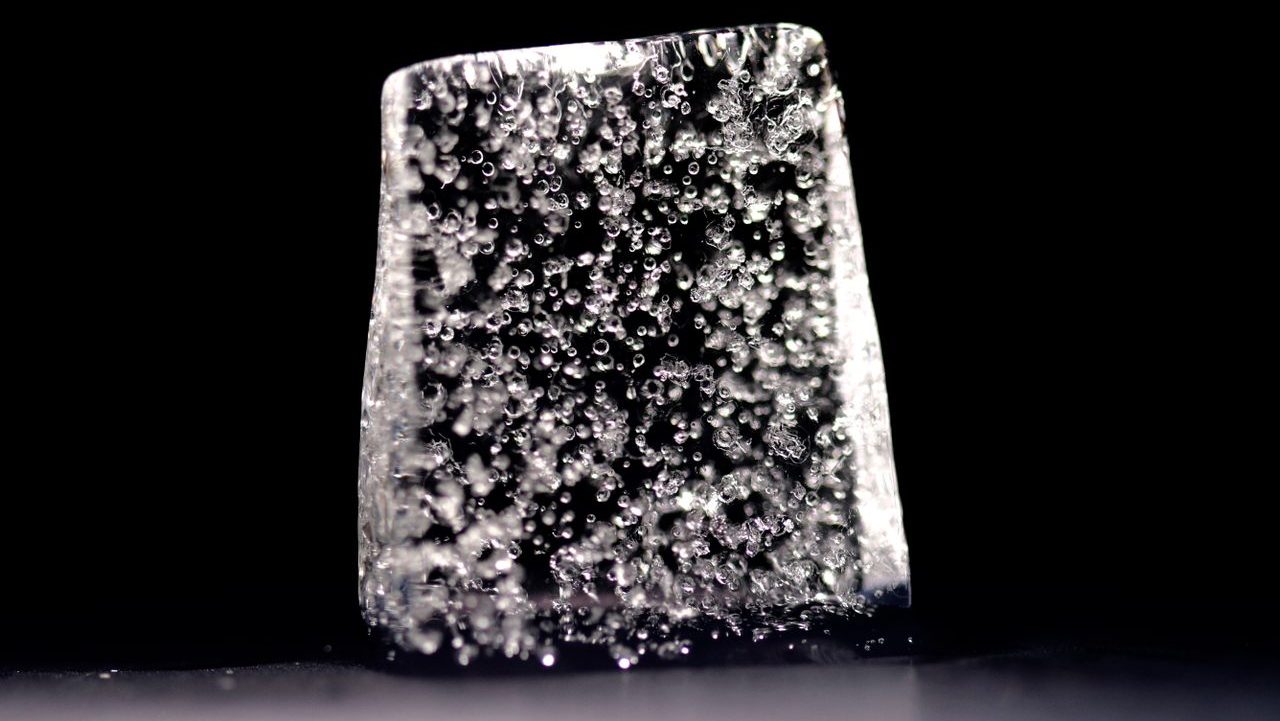 A close-up of a translucent cube of ice with a bubbly texture against a dark background.