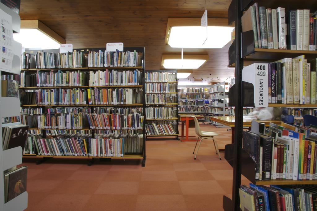 Rows of bookshelves with labeled categories in a library with wooden ceilings and pendant lighting.