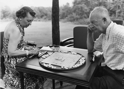 An elderly couple playing scrabble at an outdoor table, with the man watching thoughtfully as the woman considers her next move.