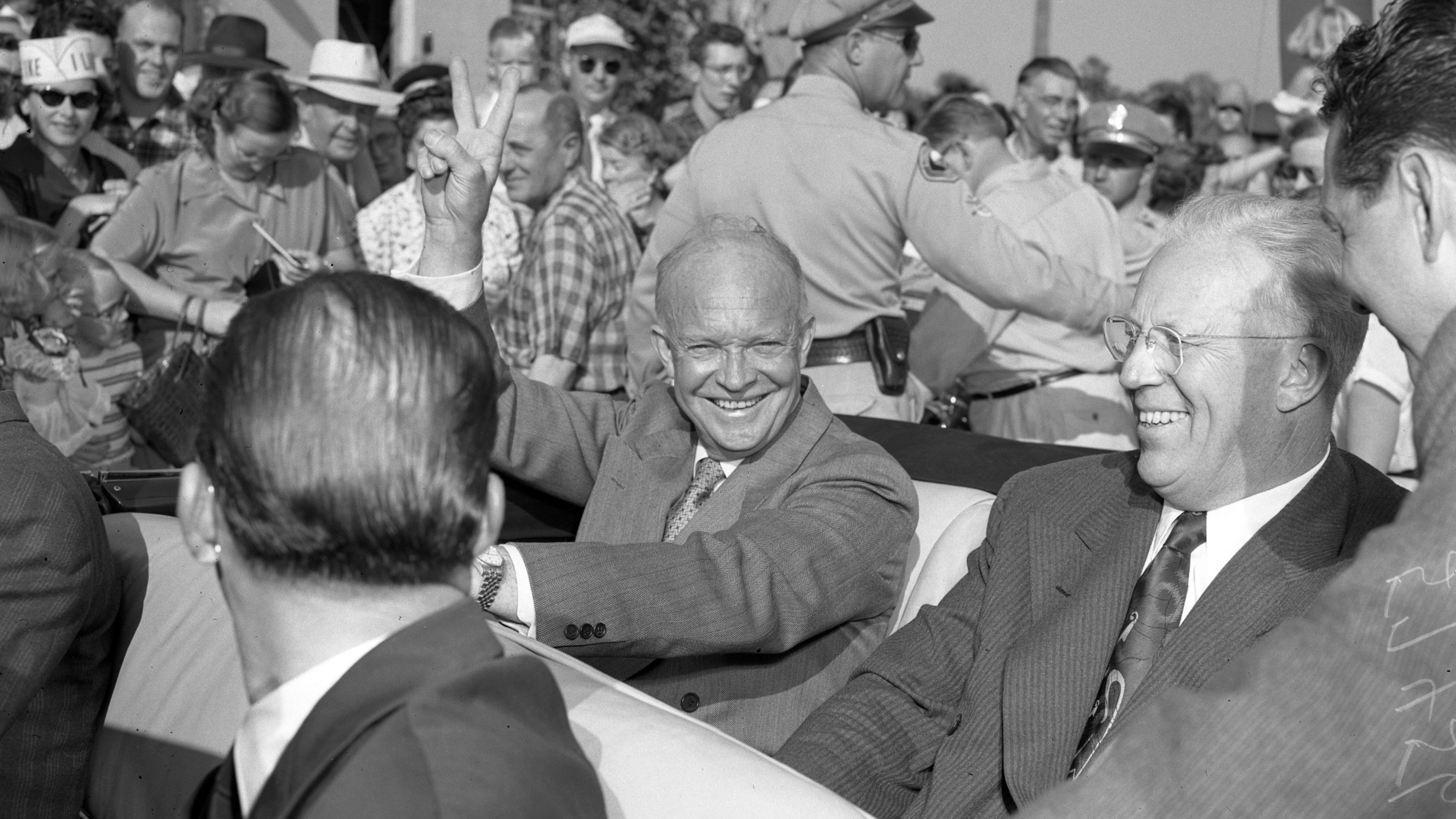 Two men smiling and riding in a convertible during a parade, surrounded by a crowd of onlookers.