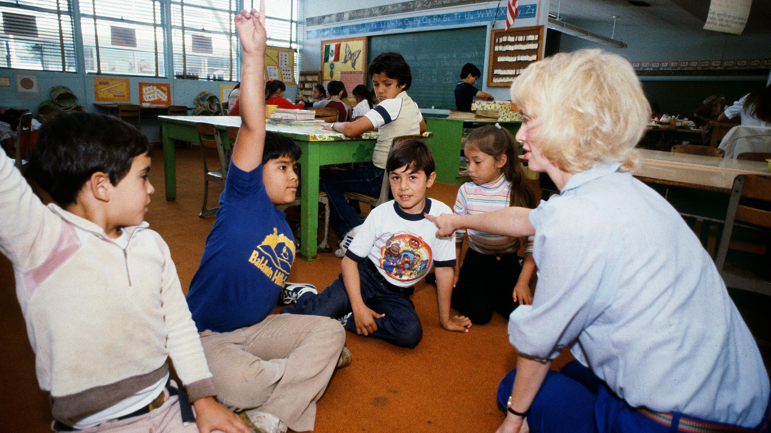 A teacher in a classroom with children sitting on the floor, learning a second language.