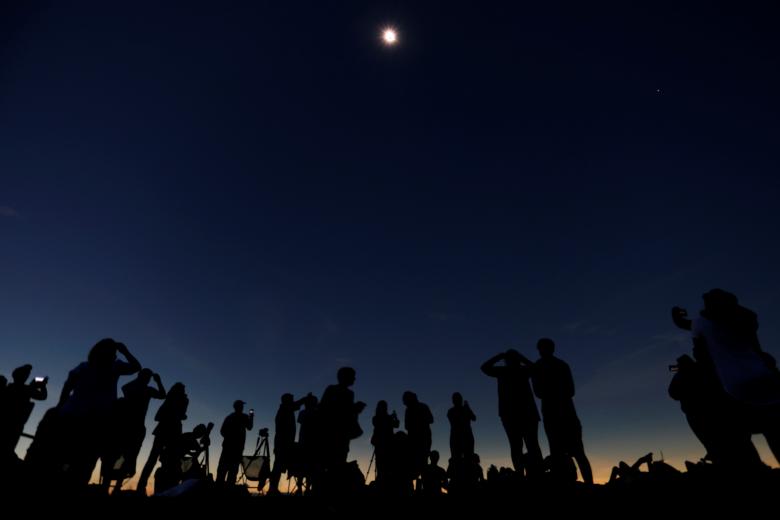 Silhouettes of people observing the total eclipse with telescopes under the moon.