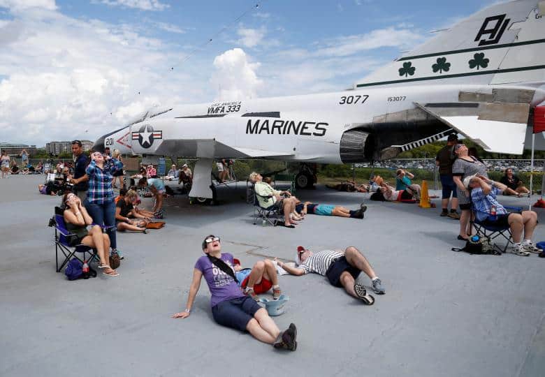 People relaxing and looking up at a total eclipse near a parked marines jet aircraft at an outdoor event.