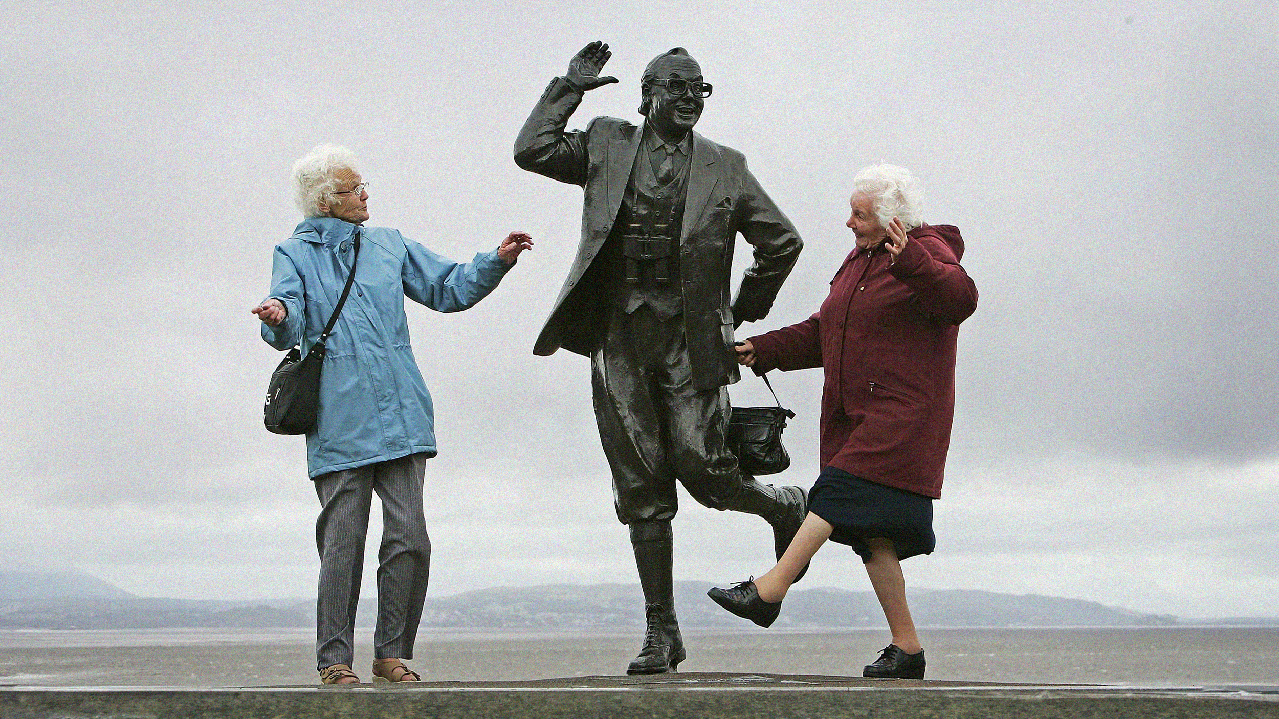 Two elderly women walking alongside and interacting with a statue of a man holding a briefcase.