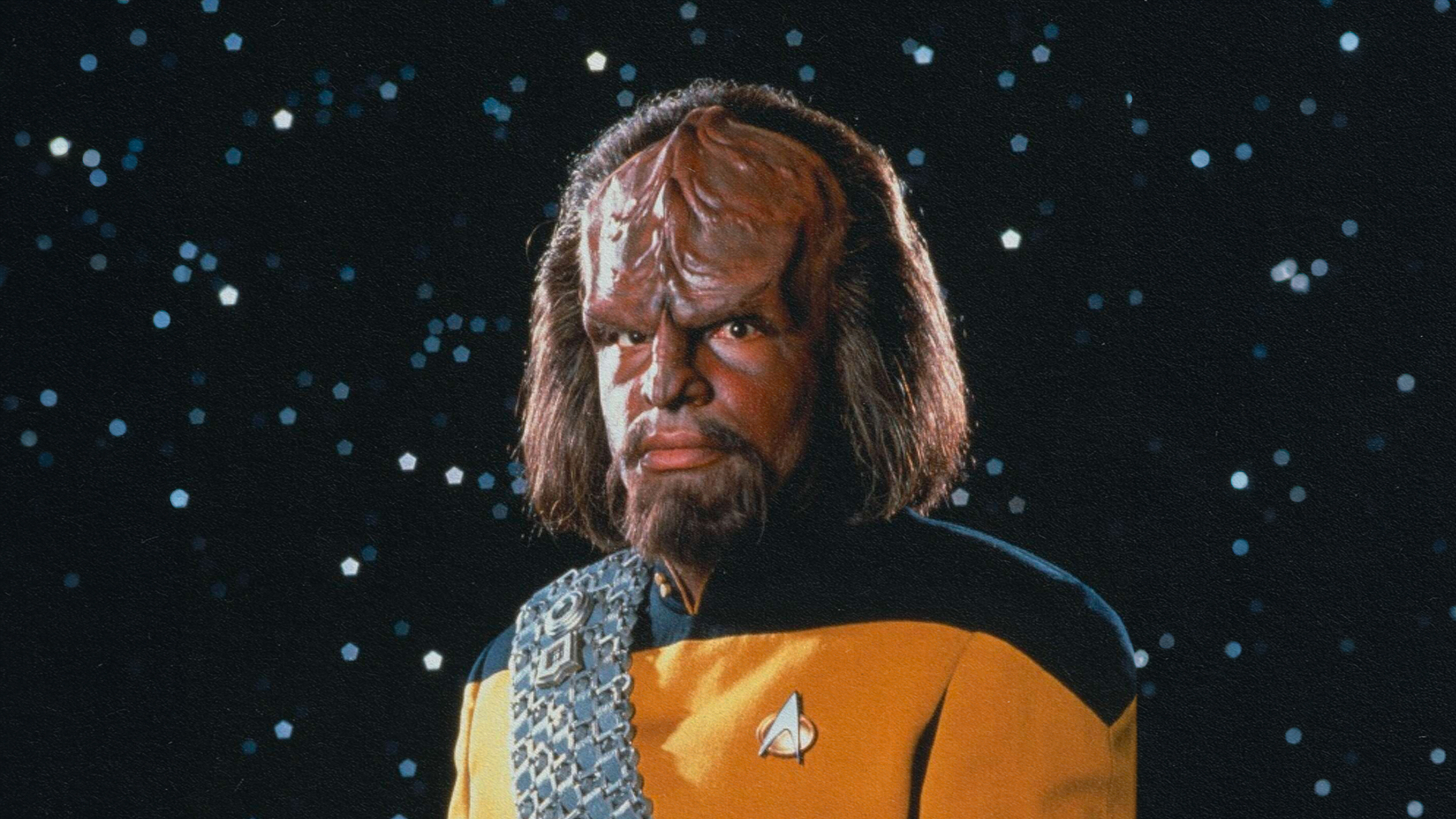 A character in a starfleet uniform with a distinctive ridged forehead, who speaks invented languages, stands against a backdrop of stars.