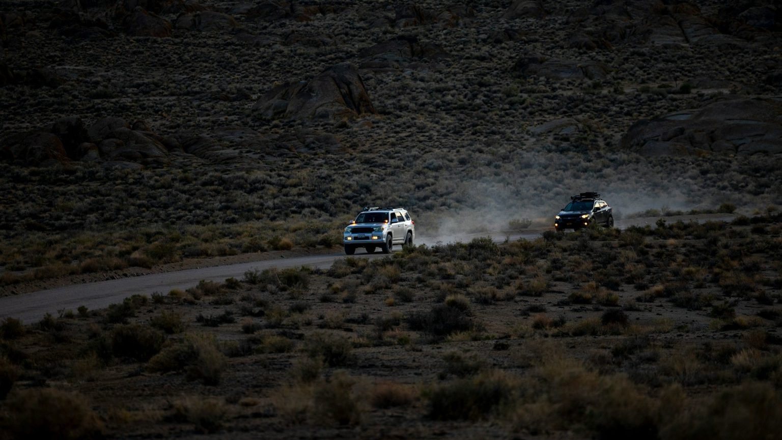 Two electric vehicles driving on a dusty road amidst desert terrain at dusk.
