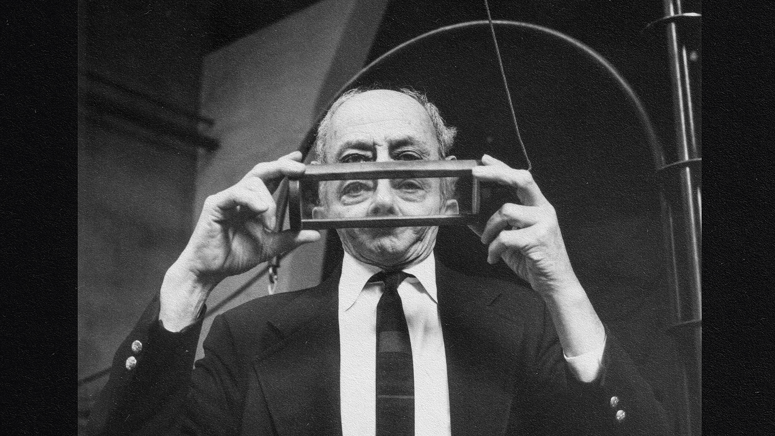 A man holding a transparent rectangular object up to his eyes, reminiscent of the analytical gaze of Robert and Frank Oppenheimer.