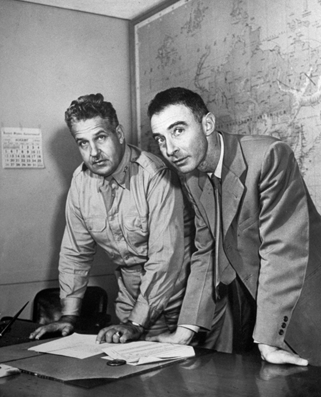 Two men in a vintage office, one leaning on a desk and the other standing, both looking towards the camera with a world map in the background.