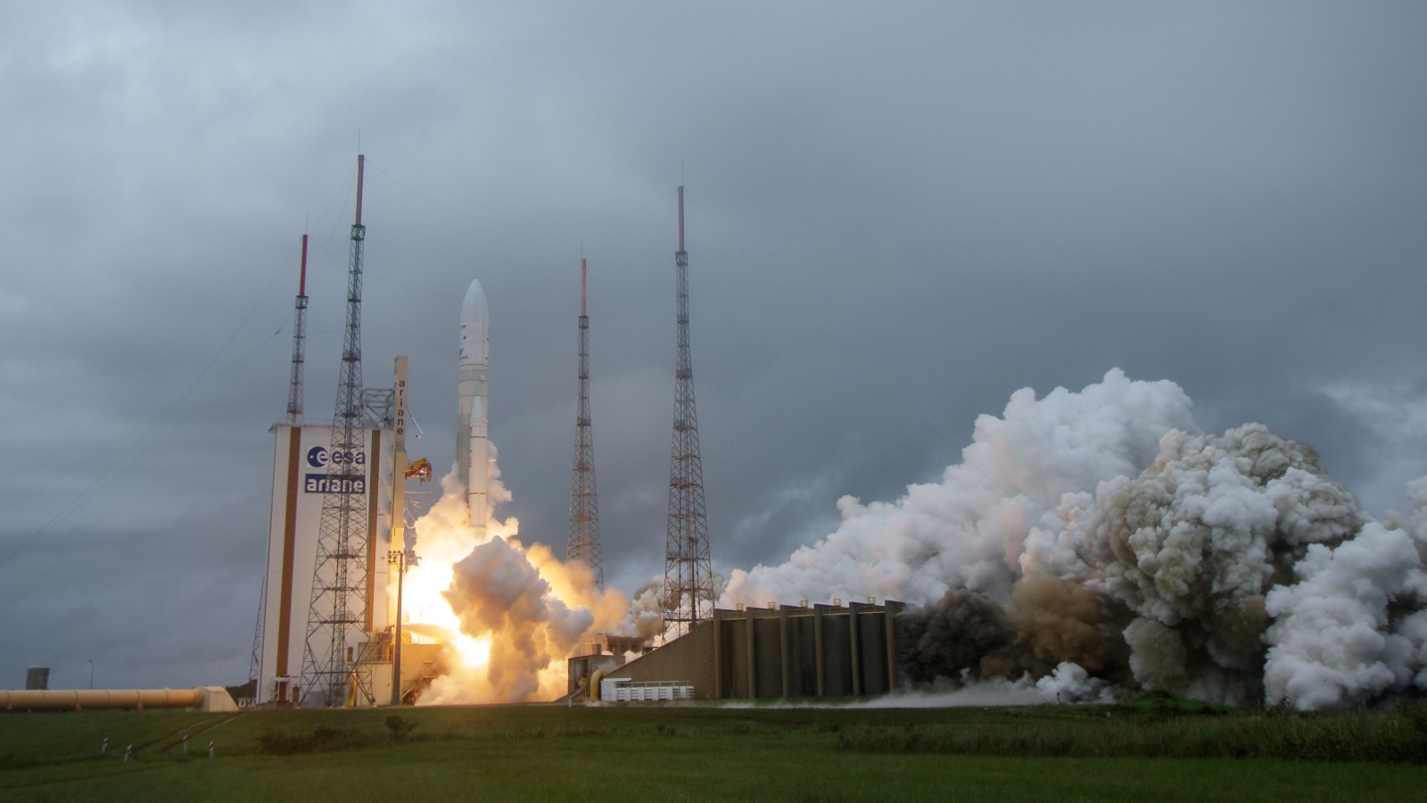 A rocket launches from its pad as smoke and steam billow out, against an overcast sky, marked with the european space agency's logo.