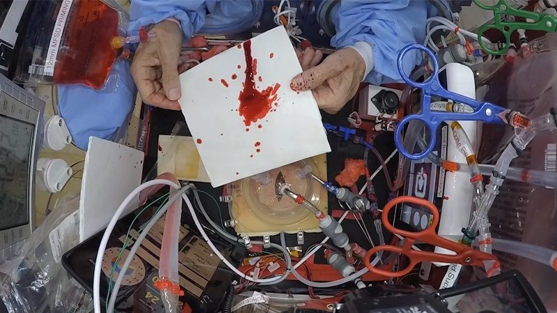 A cluttered workspace with electronic components and a hand holding a card splattered with red liquid.