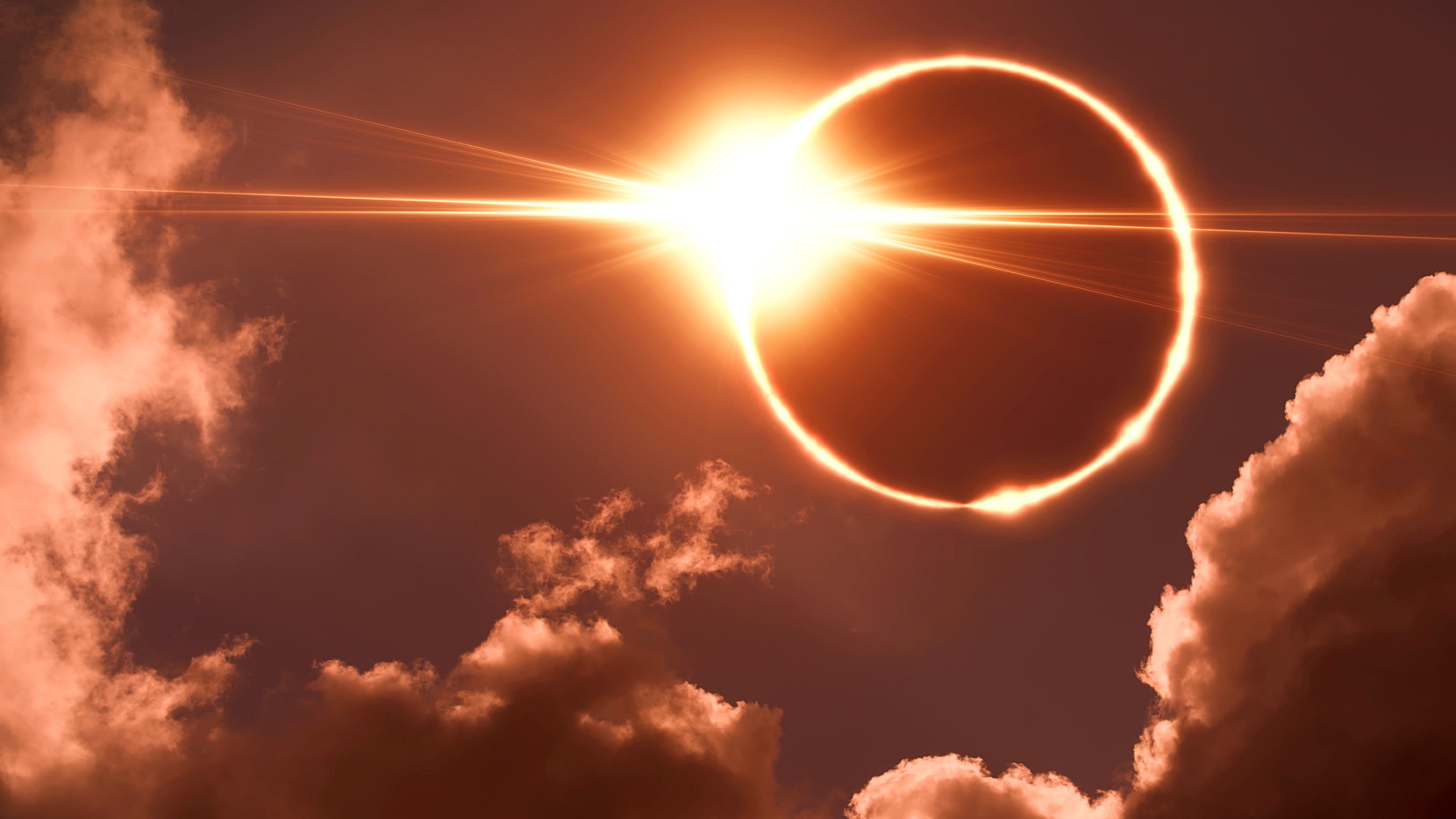 A dramatic solar eclipse with a radiant ring of fire visible around the moon, set against a backdrop of clouds and a sunburst.