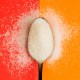 A spoonful of non-sugar sweeteners on a dual red and orange background, with sweetener grains scattered around the spoon.
