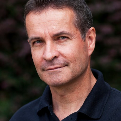 A portrait of a middle-aged man with a slight smile, wearing a black polo shirt, against a blurred natural background.