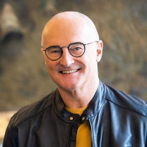 A smiling bald man with glasses, wearing a black leather jacket over a yellow sweater, in front of a blurred background.