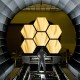 A technician in a clean suit inspects a large, segmented, hexagonal mirror inside a circular gray structure.