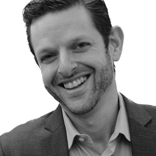 A black and white image of a smiling man with stubble, wearing a suit jacket and a collared shirt.