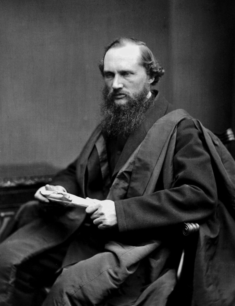 A black and white photograph of a seated man with a full beard wearing a formal coat and holding what appears to be a book or documents.
