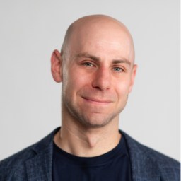 Headshot of a Adam Grant with a slight smile, wearing a dark blazer over a blue shirt against a light gray background.