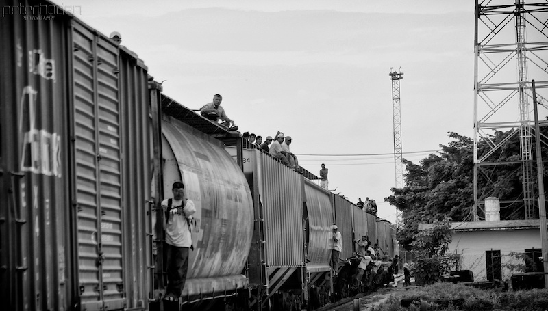 People sitting and standing on top of a moving freight train, with the Darién Gap landscape in the background.