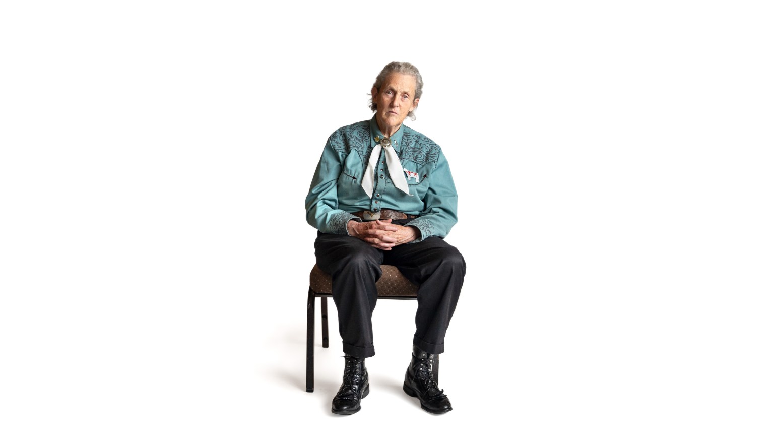 Temple Grandin sitting on a chair against a white background, dressed in a satin blouse with a scarf and black boots.