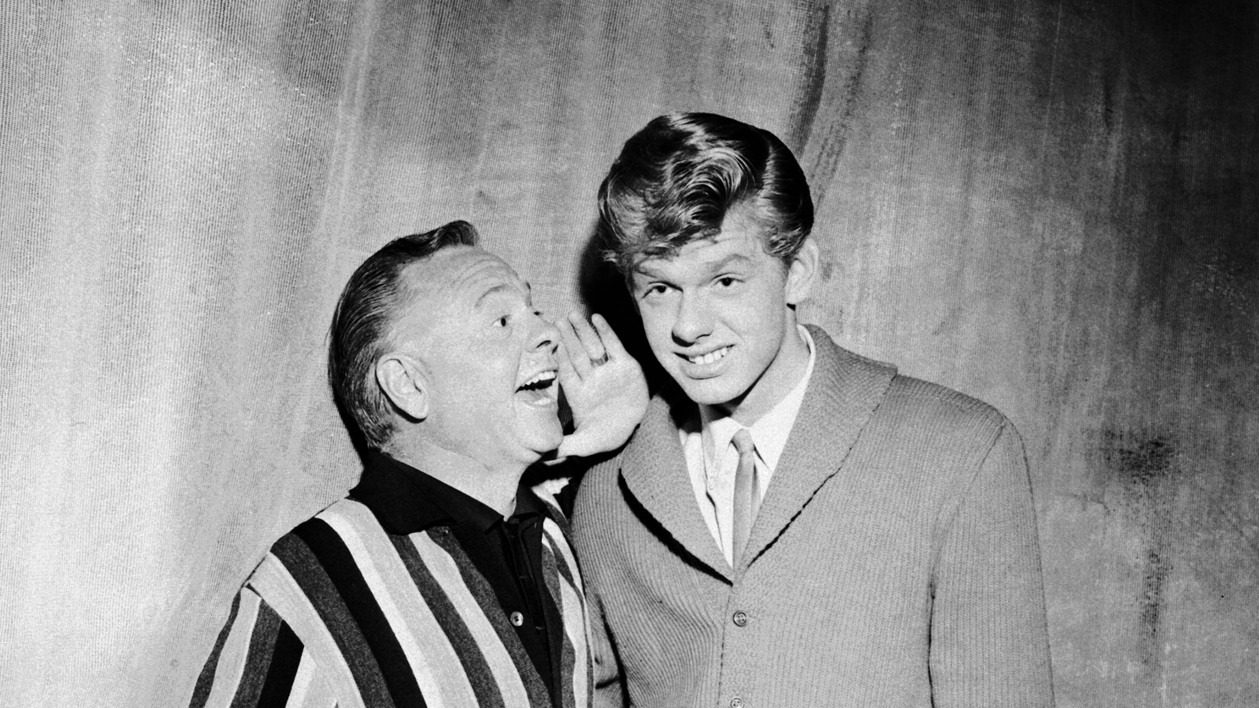 Black and white photo of an older man in a striped jacket whispering to a grinning young man in a suit.