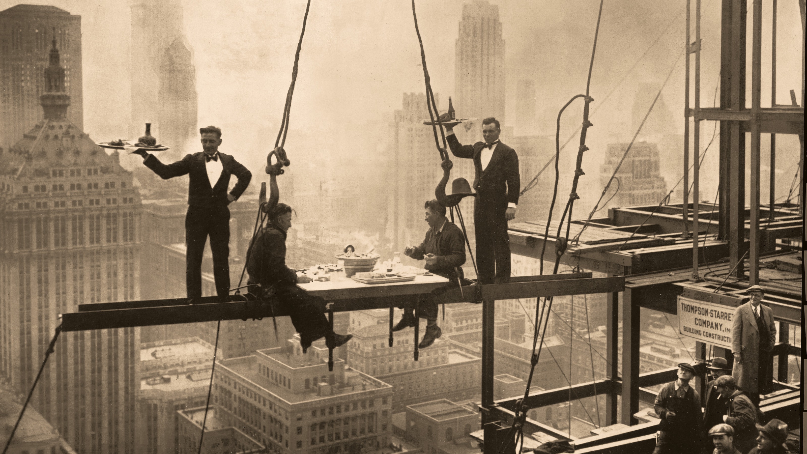 Vintage sepia-toned photo of construction workers having lunch on a steel beam high above a cityscape, with a waiter generously serving them.