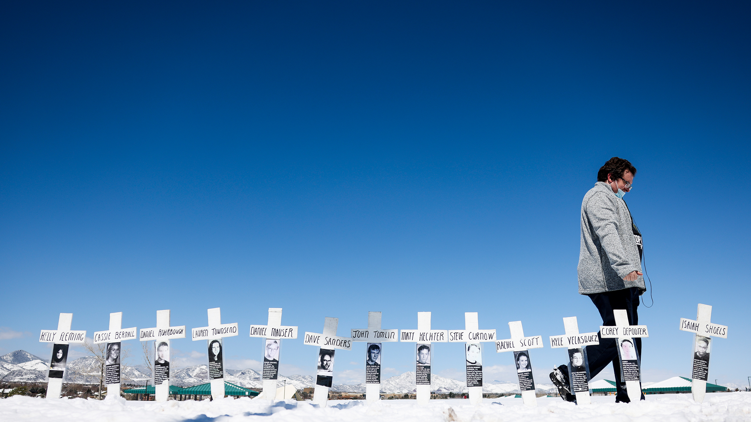 A person walking past rows of white crosses with photos and names, commemorating victims of a mass shooter, set against a snowy background and clear blue sky.