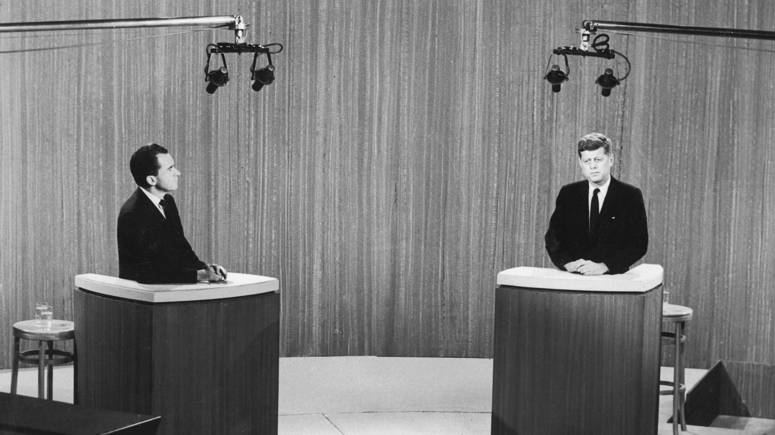 Two men from different political parties in suits stand at podiums under studio lights, participating in a televised debate.