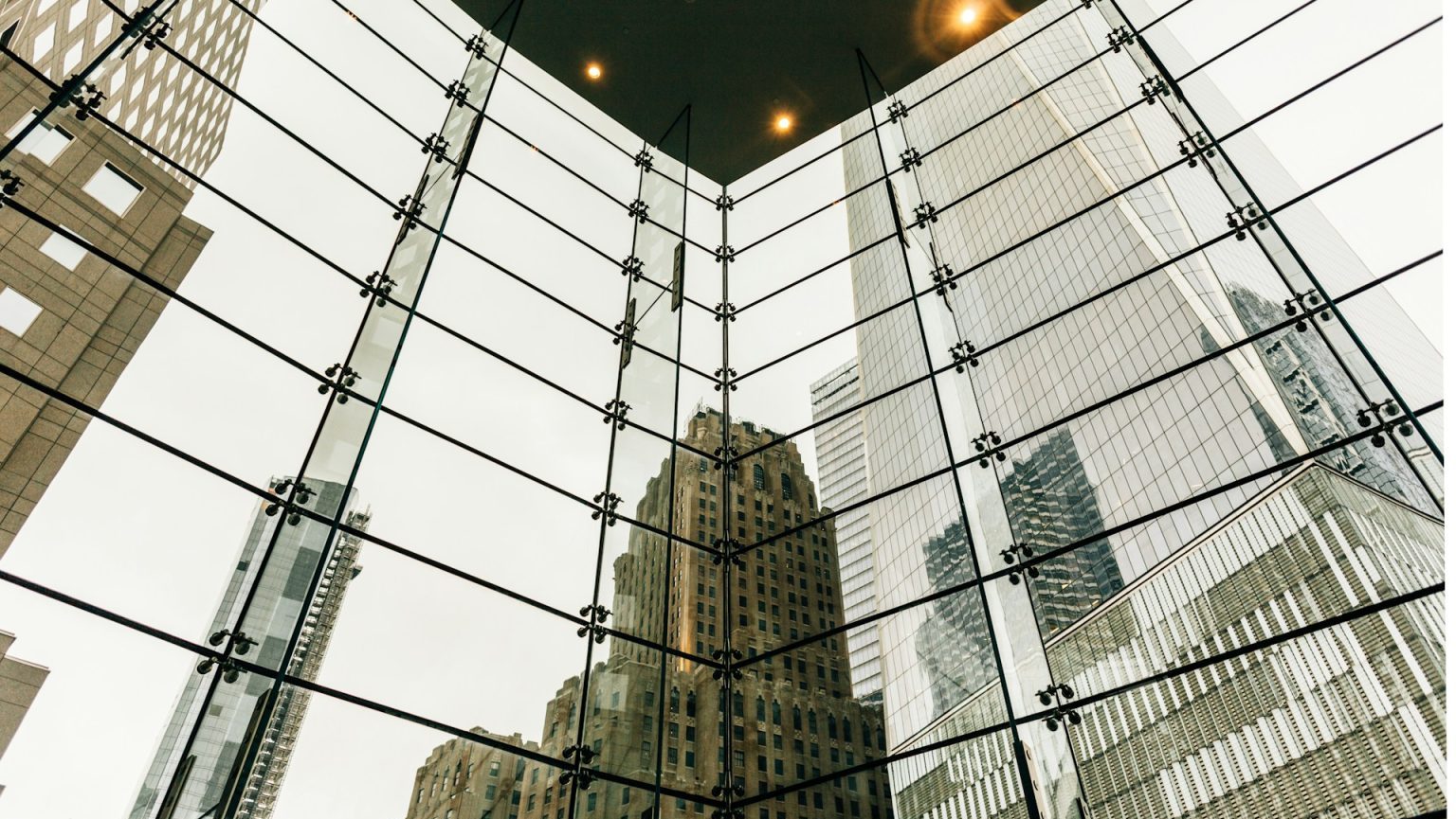 View of a cityscape through a large glass window framed by steel beams, showcasing high-rise buildings burdened with debt under an overcast sky.
