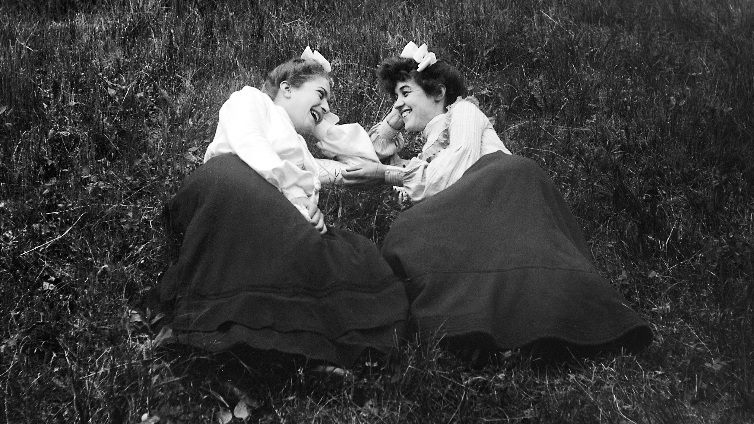 Two women in vintage clothing, wearing bows in their hair, engaging in a tend-and-befriend moment on a grassy field, lying facing each other, smiling and talking.