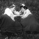 Two women in vintage clothing, wearing bows in their hair, engaging in a tend-and-befriend moment on a grassy field, lying facing each other, smiling and talking.