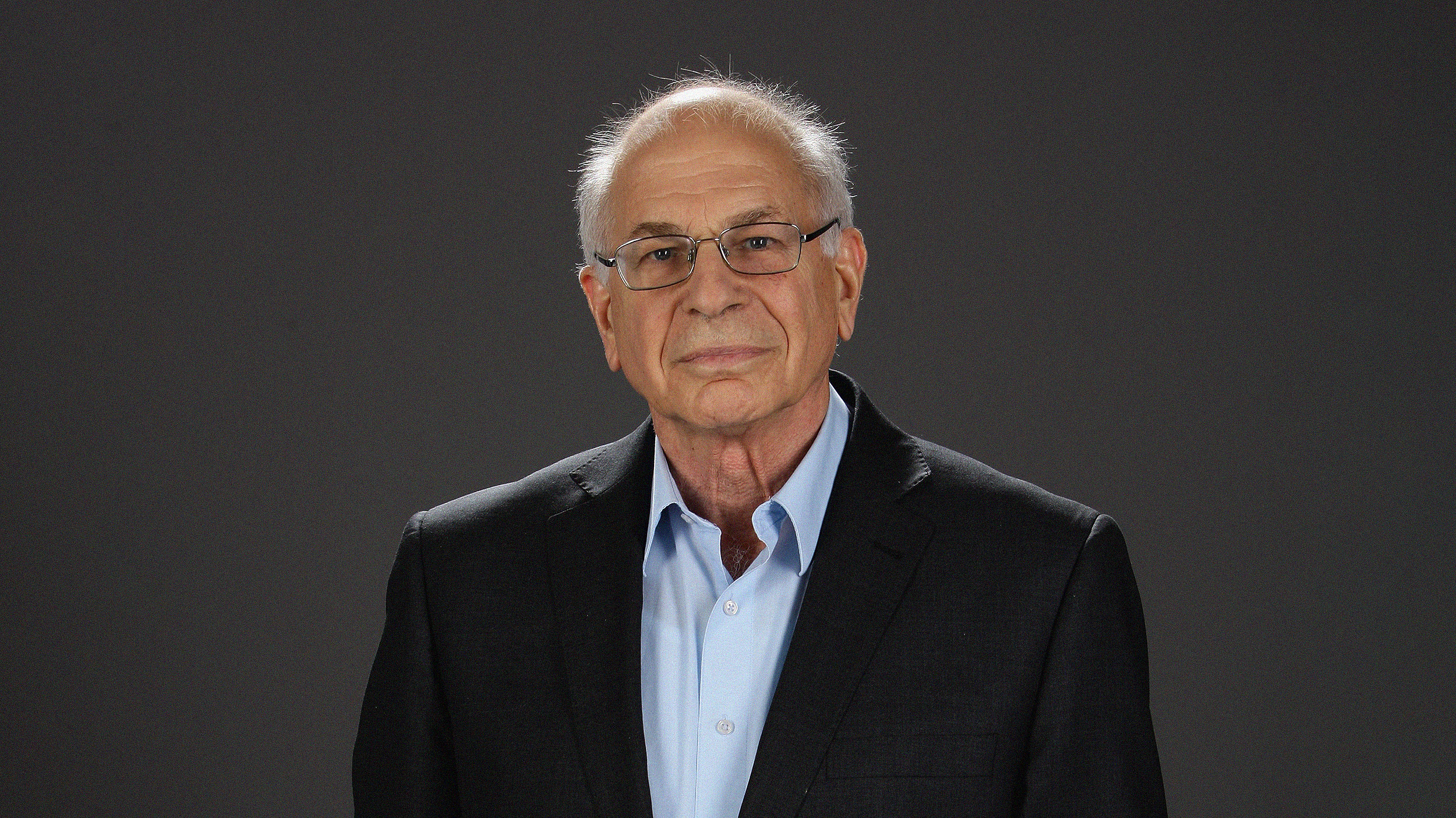 A senior man, Daniel Kahneman, with glasses, wearing a dark suit and a blue shirt, standing against a gray background.