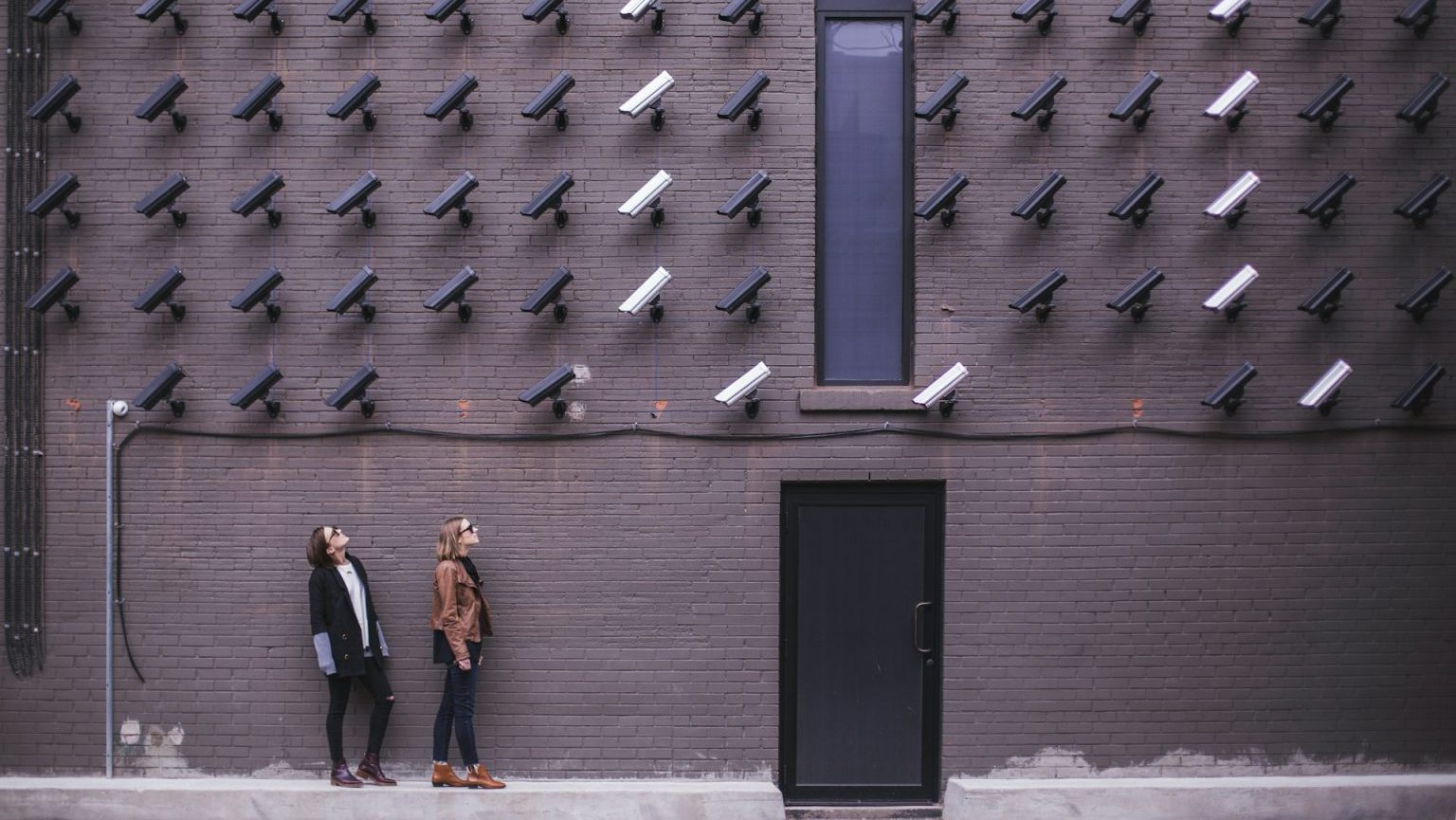 Two people walking past a dark brick wall with numerous security cameras pointing in various directions.