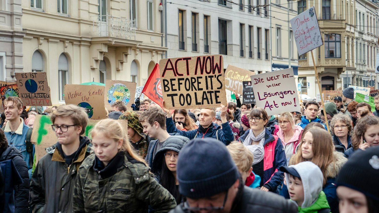 Crowd of diverse people at a climate protest holding signs with messages like "act now before it's too late".